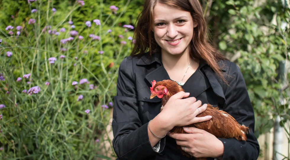Lina fra Frie Vinger holder en brun høne i favnen foran en grøn busk.